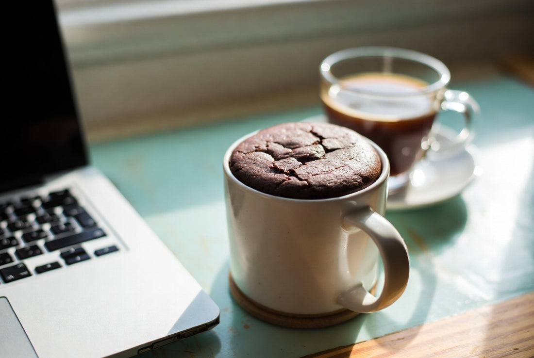 A chocolate protein mug cake beside a laptop and a cup of coffee in warm afternoon light.