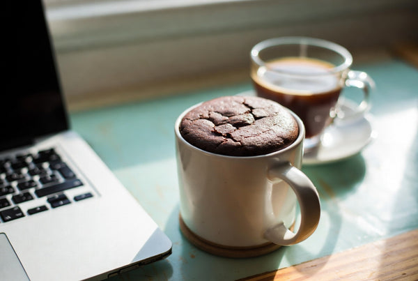 A chocolate protein mug cake beside a laptop and a cup of coffee in warm afternoon light.