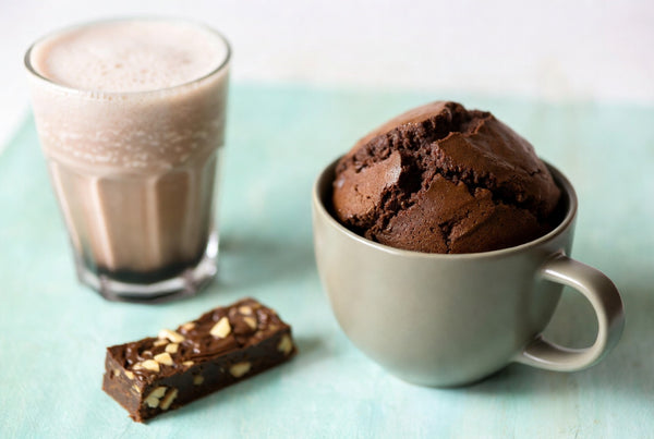 A protein shake, a protein bar, and a chocolate protein mug cake lined up side by side on a counter.